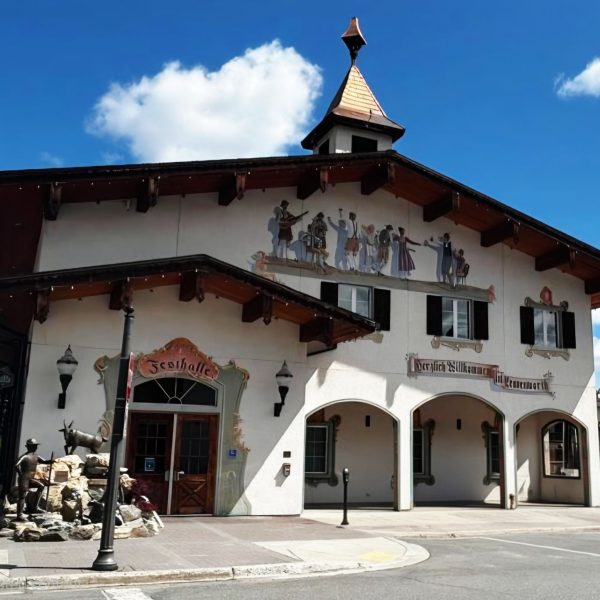 A Bavarian-style building with a steep roof, mural of dancers above, and a small clock tower. A stone fountain with animal sculptures stands by the entrance. The sky is blue with scattered clouds.