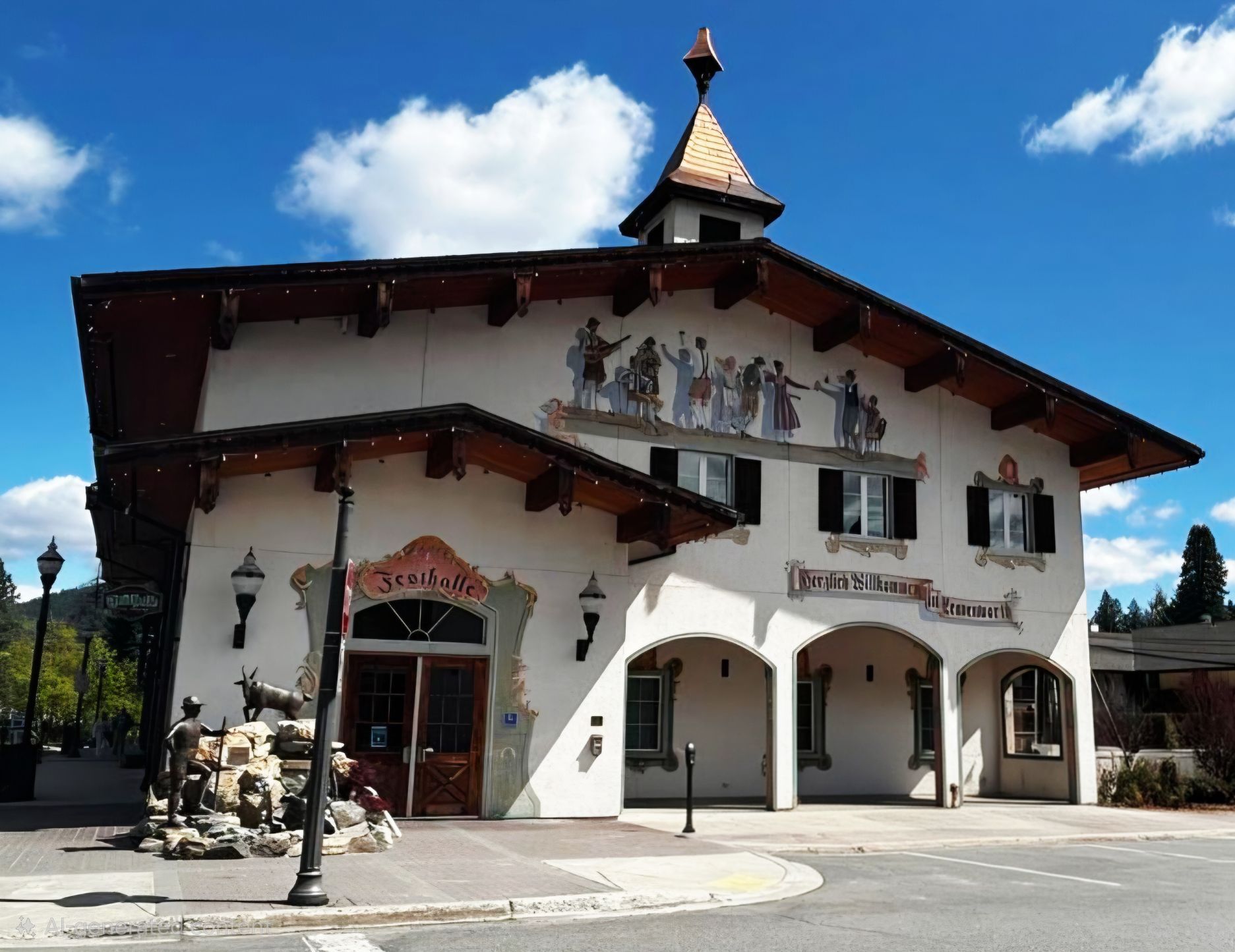 A Bavarian-style building with a steep roof, mural of dancers above, and a small clock tower. A stone fountain with animal sculptures stands by the entrance. The sky is blue with scattered clouds.