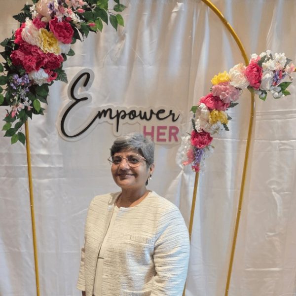 A woman with short gray hair and glasses stands smiling in front of a floral arch and a backdrop that reads Empower HER. She is wearing a light-colored jacket.