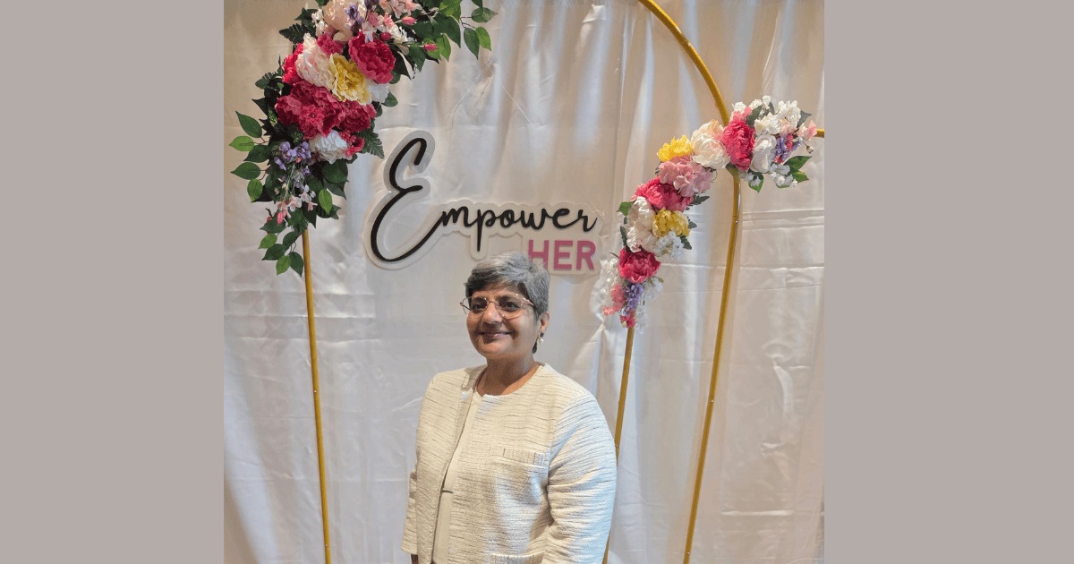 A woman with short gray hair and glasses stands smiling in front of a floral arch and a backdrop that reads Empower HER. She is wearing a light-colored jacket.