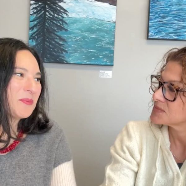 Two women sitting at a table indoors, both wearing glasses and smiling. The background features an art-decorated wall.