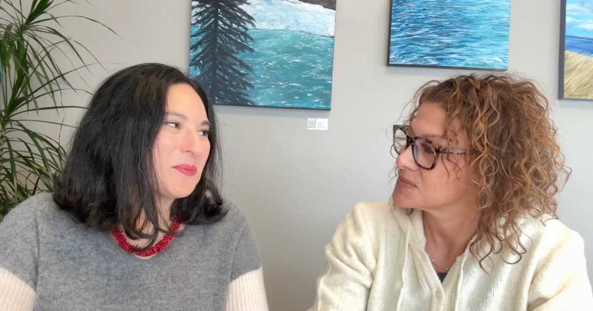 Two women sitting at a table indoors, both wearing glasses and smiling. The background features an art-decorated wall.