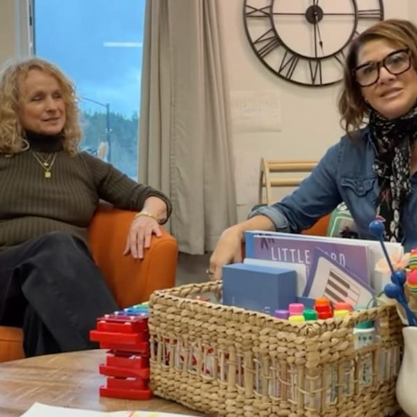 Two women sitting in an orange armchair, one with curly blonde hair and the other wearing glasses, next to a table with colorful blocks and a decorative basket. A clock is visible on the wall behind them.