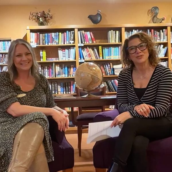 Two women sitting in purple chairs, smiling, with a globe on a table between them in front of bookshelves in a cozy library or lounge setting. One holds papers, the other has hands on her knees.