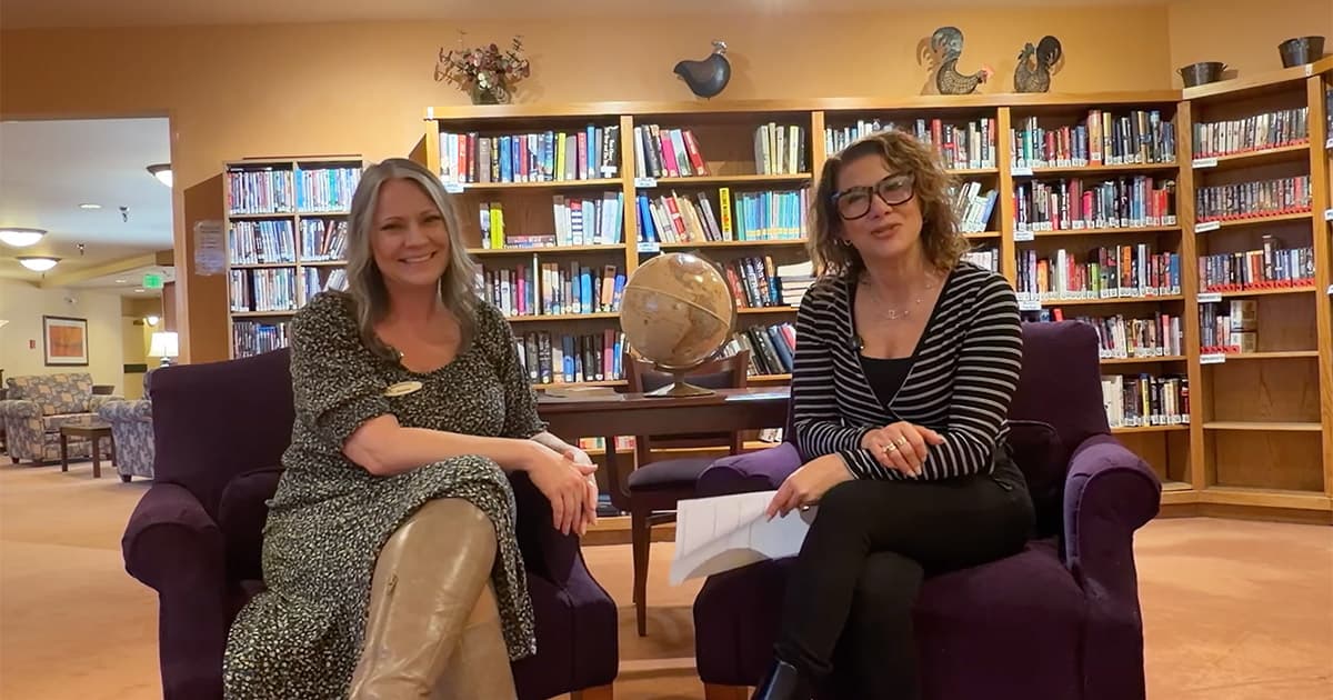 Two women sitting in purple chairs, smiling, with a globe on a table between them in front of bookshelves in a cozy library or lounge setting. One holds papers, the other has hands on her knees.