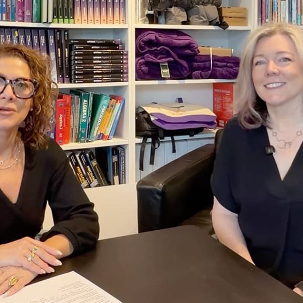 Two women sit at a black table in an office or library setting, smiling at the camera. Bookshelves filled with books, purple blankets, and boxes are in the background. Both women are wearing black tops.