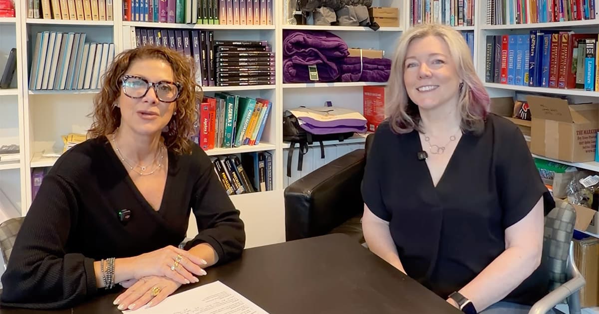 Two women sit at a black table in an office or library setting, smiling at the camera. Bookshelves filled with books, purple blankets, and boxes are in the background. Both women are wearing black tops.
