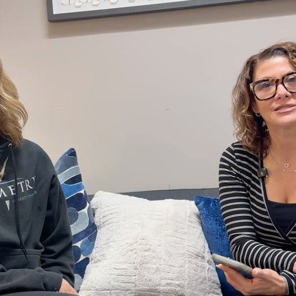 Two women sit on a couch with pillows, one wearing a black hoodie and smiling, the other wearing glasses and a black striped top, holding a phone and looking toward the camera.