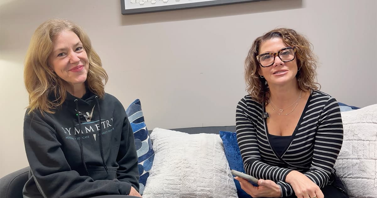 Two women sit on a couch with pillows, one wearing a black hoodie and smiling, the other wearing glasses and a black striped top, holding a phone and looking toward the camera.