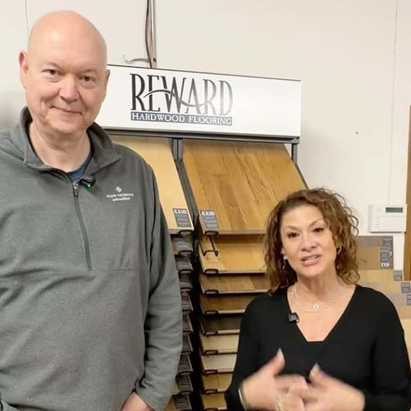 A tall man and a woman stand in front of wood flooring samples in a showroom, with a Reward Hardwood Flooring sign above them. The man wears a gray pullover and the woman is dressed in black.