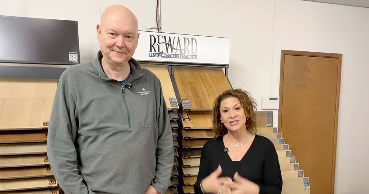 A tall man and a woman stand in front of wood flooring samples in a showroom, with a Reward Hardwood Flooring sign above them. The man wears a gray pullover and the woman is dressed in black.