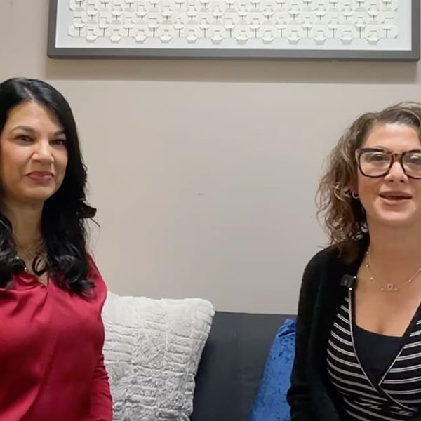 Two women are sitting on a couch in a living room, facing the camera and smiling. One has long dark hair and wears a red top; the other has curly hair, glasses, and wears a black cardigan over a striped shirt.