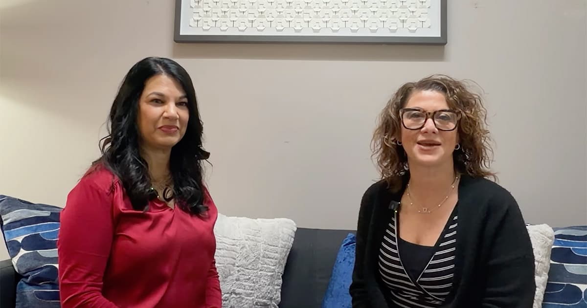 Two women are sitting on a couch in a living room, facing the camera and smiling. One has long dark hair and wears a red top; the other has curly hair, glasses, and wears a black cardigan over a striped shirt.