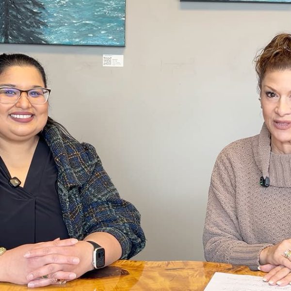 Two women sit at a wooden table, smiling at the camera. One wears glasses and a plaid jacket, the other wears a tan sweater. Artworks and a plant are visible in the background.