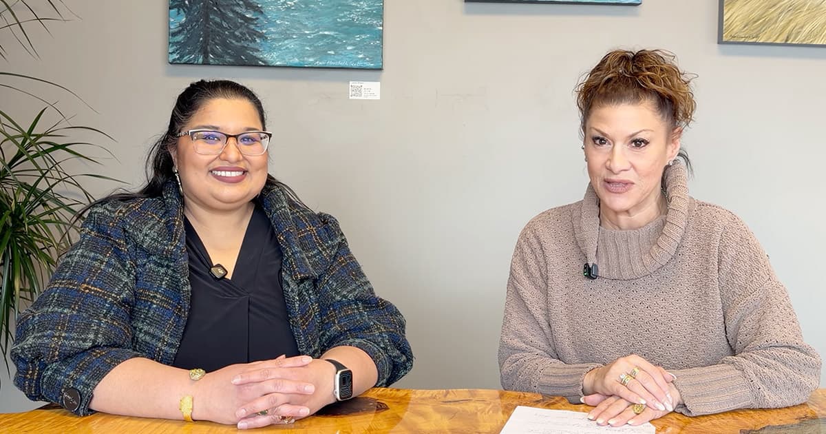 Two women sit at a wooden table, smiling at the camera. One wears glasses and a plaid jacket, the other wears a tan sweater. Artworks and a plant are visible in the background.