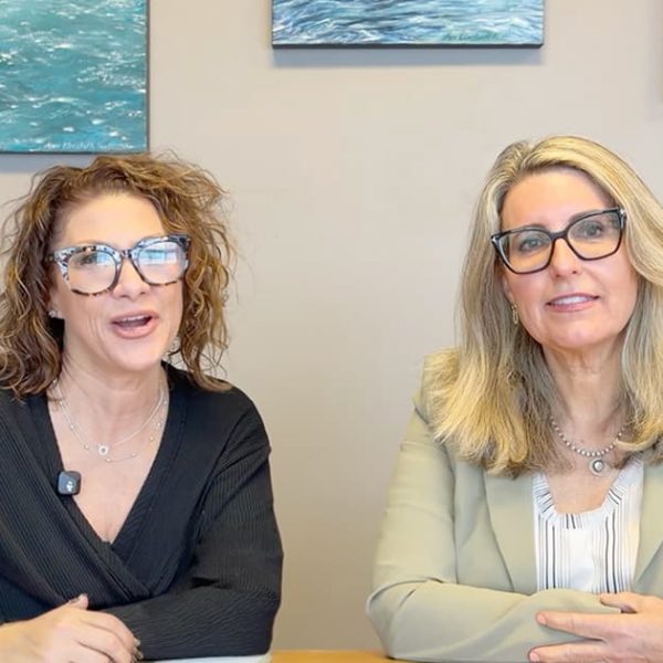Two women with light hair sit at a table, smiling at the camera. Both wear glasses and microphones. Behind them are paintings of nature on a beige wall.