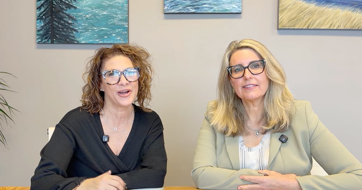Two women with light hair sit at a table, smiling at the camera. Both wear glasses and microphones. Behind them are paintings of nature on a beige wall.