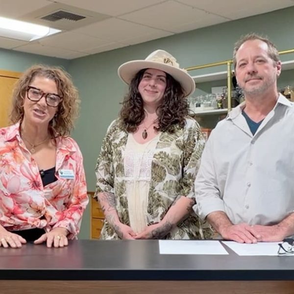 Three adults stand behind a counter in a natural health store. The woman on the left wears glasses and a floral blouse, the woman in the middle wears a hat, and the man on the right wears a light shirt. Plants and products are visible.