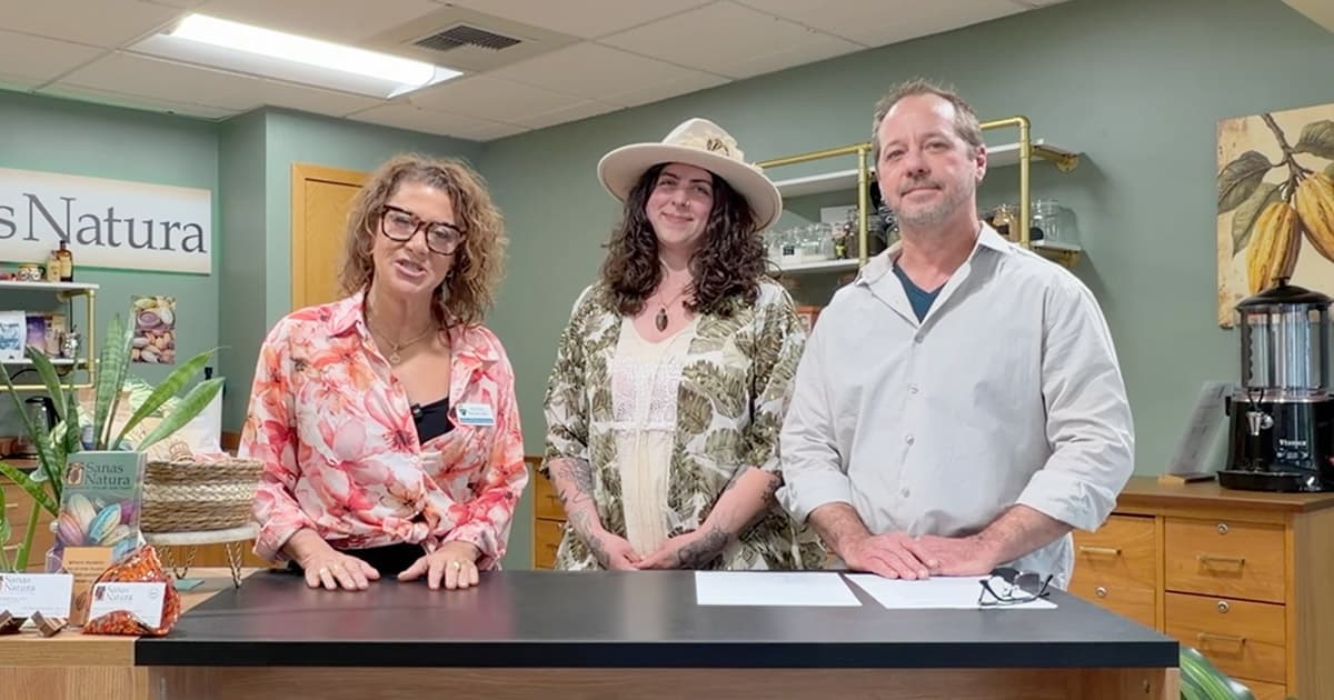 Three adults stand behind a counter in a natural health store. The woman on the left wears glasses and a floral blouse, the woman in the middle wears a hat, and the man on the right wears a light shirt. Plants and products are visible.