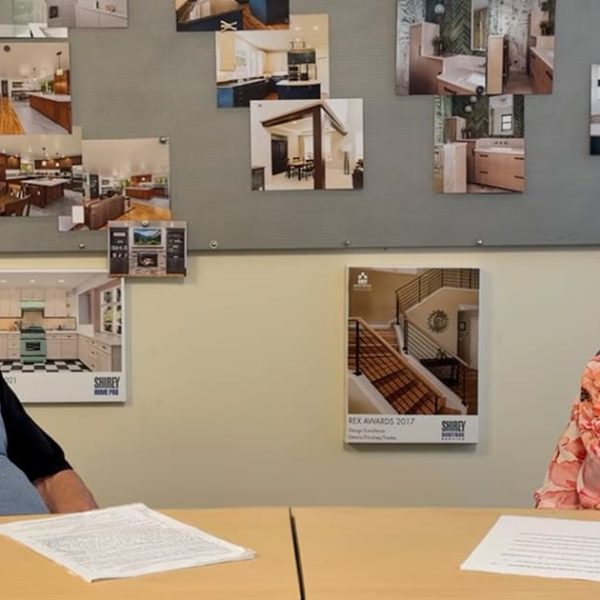 Two people sit at a table with papers in front of them, talking. Behind them, a bulletin board displays photos of home interiors and remodeling projects. Both appear engaged in conversation.