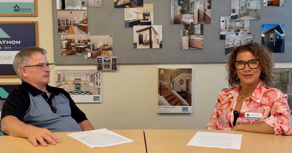 Two people sit at a table with papers in front of them, talking. Behind them, a bulletin board displays photos of home interiors and remodeling projects. Both appear engaged in conversation.