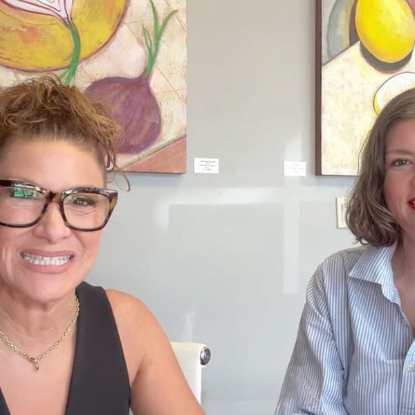 Two women smiling and sitting at a table, with colorful fruit-themed artwork hanging on the wall behind them. One woman wears glasses and a black top; the other has shoulder-length hair and a light blue shirt.