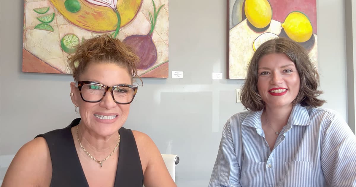 Two women smiling and sitting at a table, with colorful fruit-themed artwork hanging on the wall behind them. One woman wears glasses and a black top; the other has shoulder-length hair and a light blue shirt.