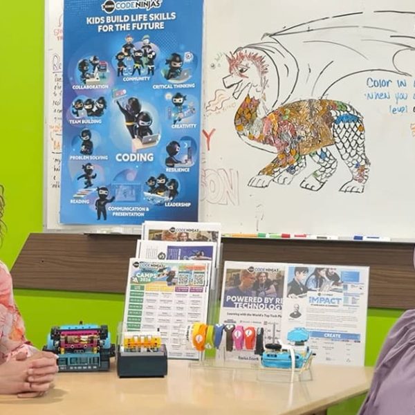 Two women sit at a table in a colorful classroom with coding posters, robotics kits, and a dragon drawing on a whiteboard behind them. The atmosphere is educational and welcoming.