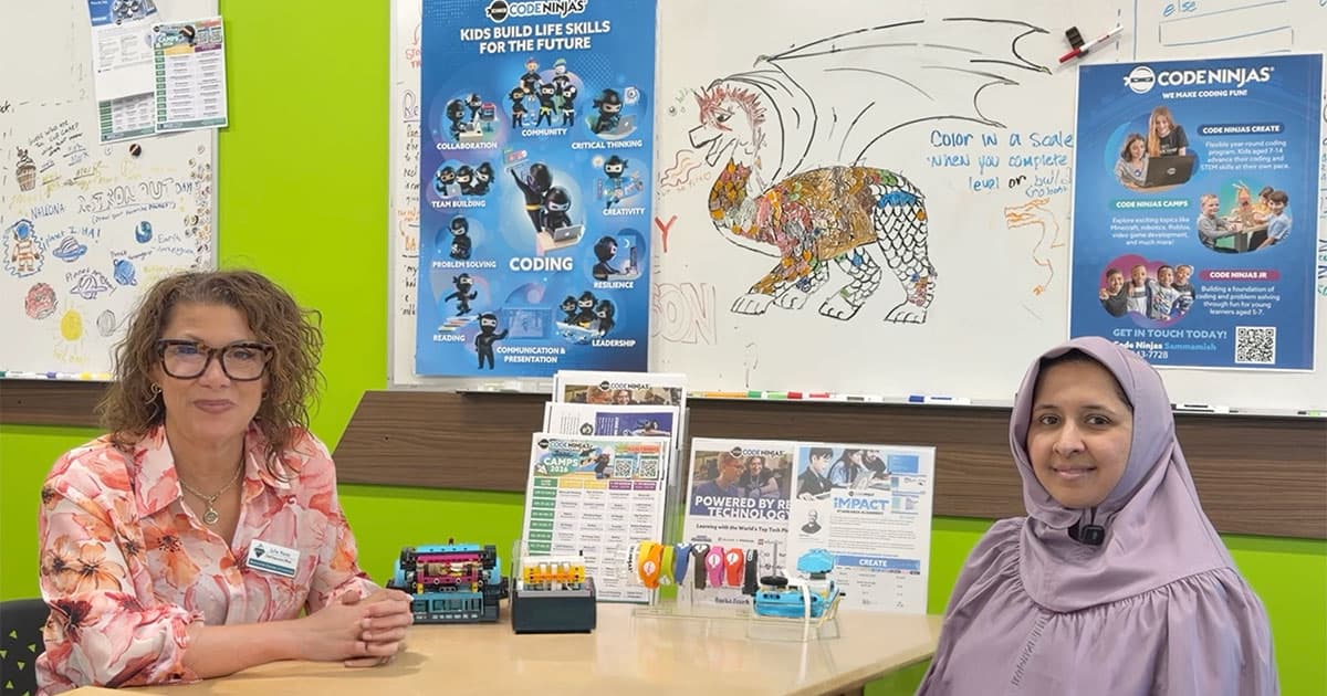 Two women sit at a table in a colorful classroom with coding posters, robotics kits, and a dragon drawing on a whiteboard behind them. The atmosphere is educational and welcoming.
