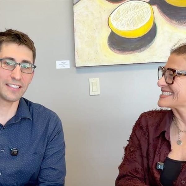 Two people wearing glasses sit and smile at each other in front of colorful artwork featuring lemons and other shapes on a light gray wall. Both are wearing microphones clipped to their shirts.