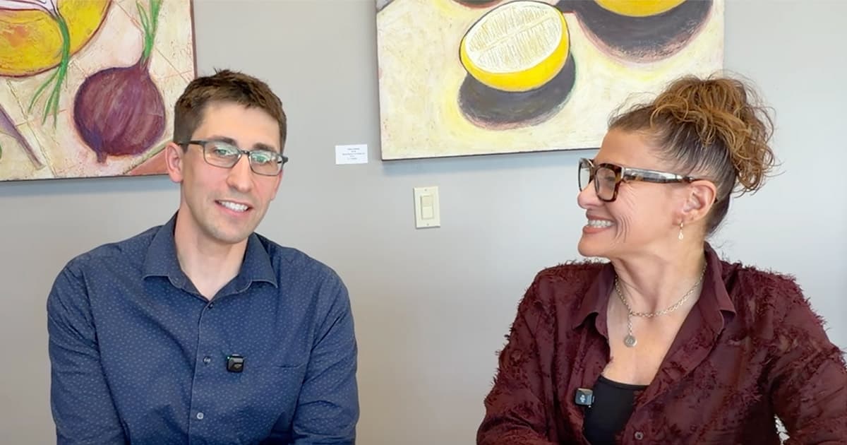 Two people wearing glasses sit and smile at each other in front of colorful artwork featuring lemons and other shapes on a light gray wall. Both are wearing microphones clipped to their shirts.