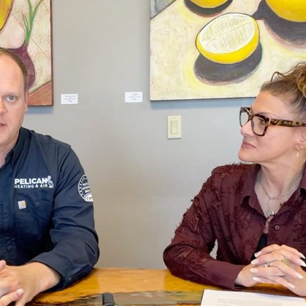 Two people sit at a table in front of colorful fruit paintings. The man wears a dark shirt with Pelican on it, and the woman wears glasses and a maroon blouse, looking at him while he faces the camera.