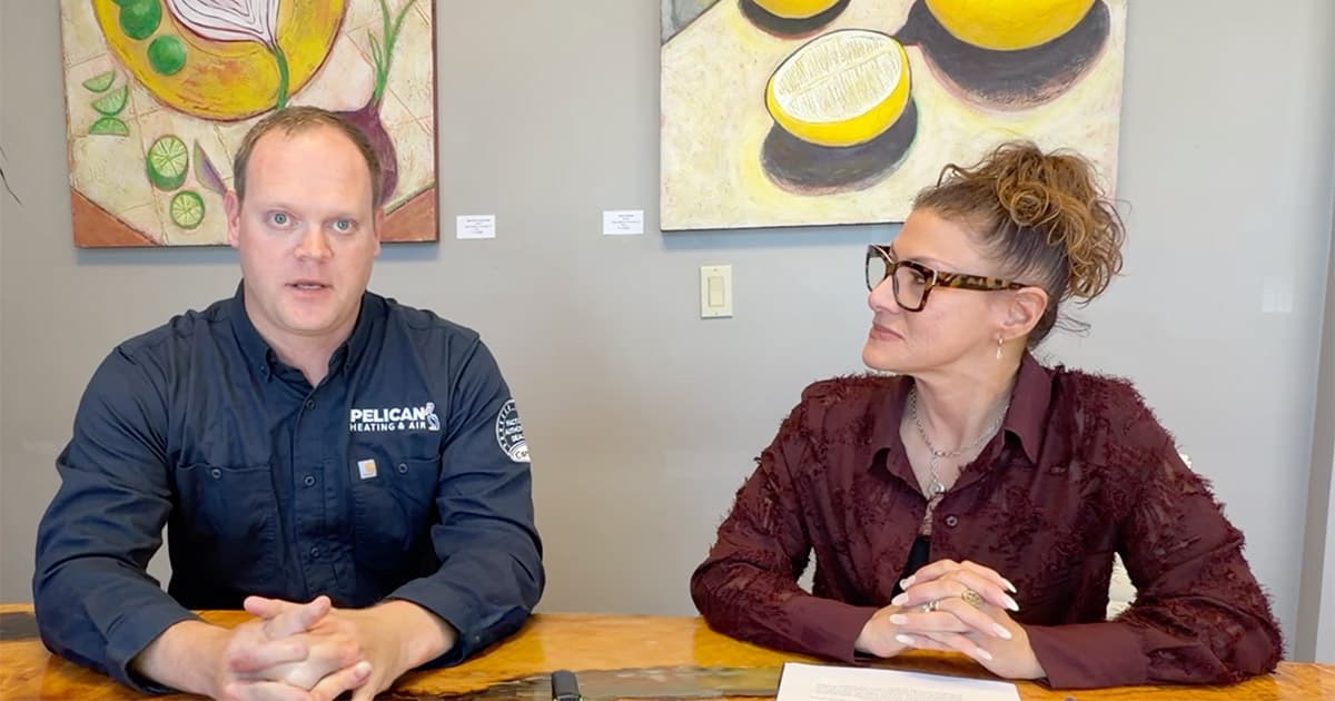 Two people sit at a table in front of colorful fruit paintings. The man wears a dark shirt with Pelican on it, and the woman wears glasses and a maroon blouse, looking at him while he faces the camera.