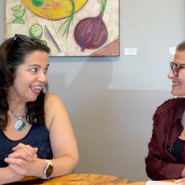 Two women sit at a wooden table, smiling and talking. One wears a dark sleeveless top, the other wears glasses and a maroon shirt. Colorful fruit paintings hang on the wall behind them.