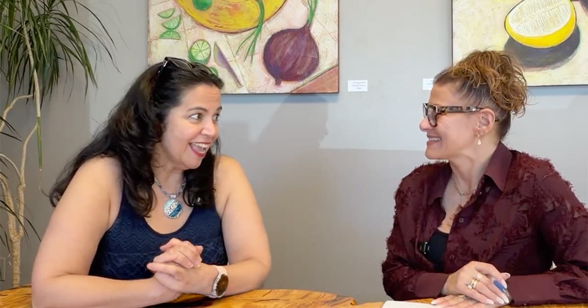 Two women sit at a wooden table, smiling and talking. One wears a dark sleeveless top, the other wears glasses and a maroon shirt. Colorful fruit paintings hang on the wall behind them.