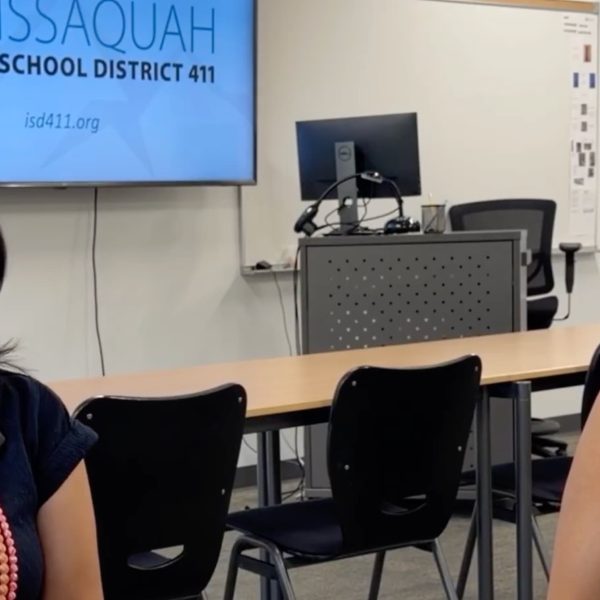 Two women sit in a meeting room with an Issaquah School District logo displayed on a wall monitor behind them.