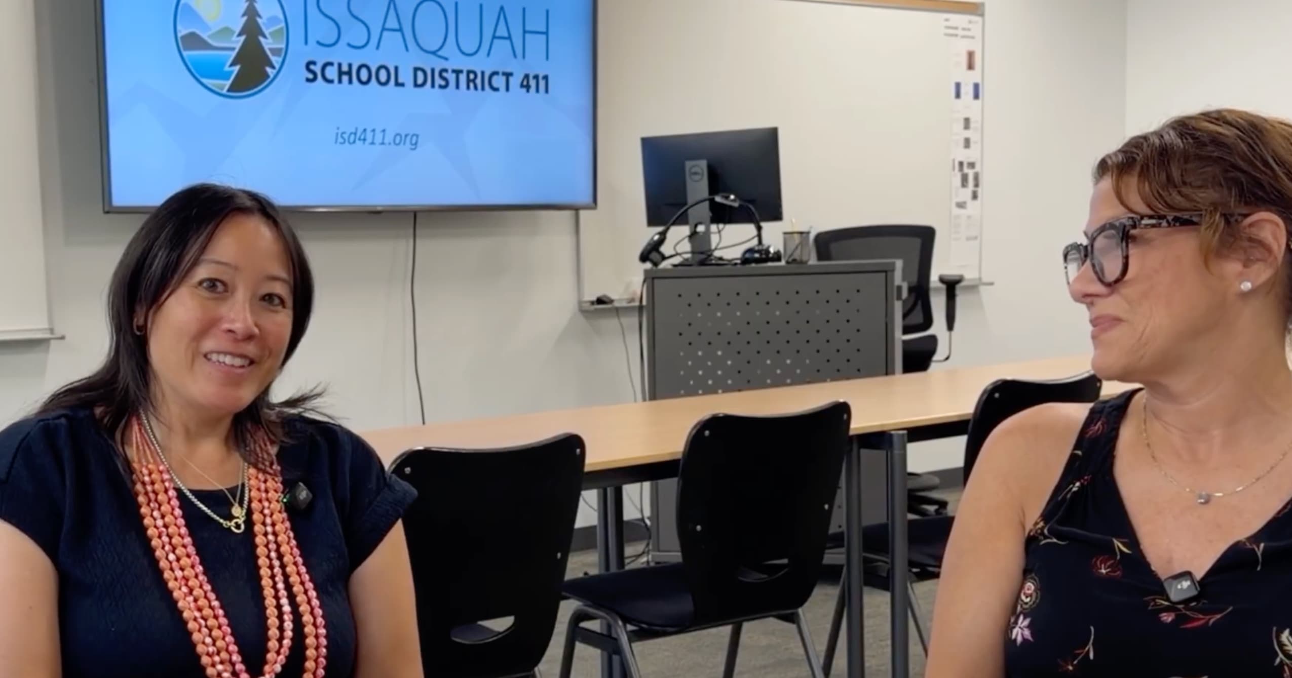 Two women sit in a meeting room with an Issaquah School District logo displayed on a wall monitor behind them.