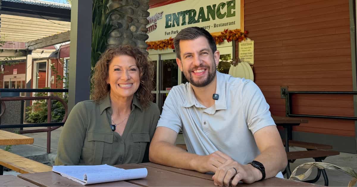 A smiling man and woman sitting at an outdoor picnic table in front of a restaurant entrance.