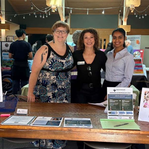 Three women stand smiling behind a booth at a nonprofit expo. The table displays informational materials and a sign reading “Connect for Good.” Other booths and attendees are visible in the background.