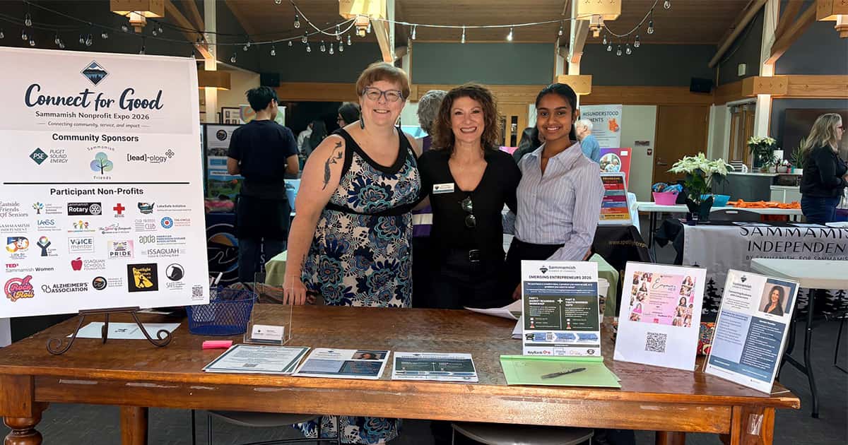 Three women stand smiling behind a booth at a nonprofit expo. The table displays informational materials and a sign reading “Connect for Good.” Other booths and attendees are visible in the background.
