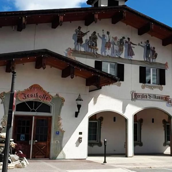 A white building with Alpine-style architecture, wooden eaves, and arched entryways, features painted murals of dancers and musicians, and a small fountain with deer statues in front. Blue sky and trees are in the background.
