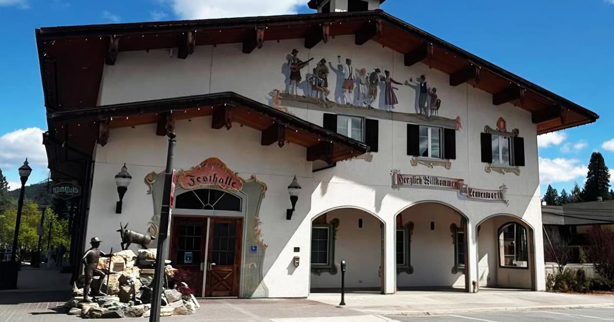 A white building with Alpine-style architecture, wooden eaves, and arched entryways, features painted murals of dancers and musicians, and a small fountain with deer statues in front. Blue sky and trees are in the background.